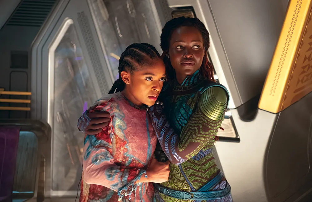 A scene from Black Panther: Wakanda Forever featuring Nakia embracing young Prince Toussaint inside a futuristic Wakandan setting. Warm cinematic lighting illuminates the pair as they stand together amid the advanced metallic architecture, emphasizing the emotional bond between mother and son.