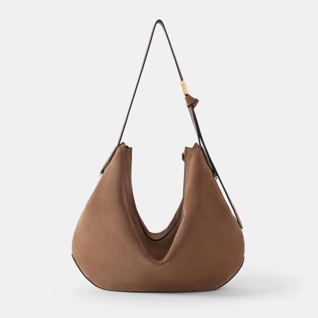 Soft brown suede hobo shoulder bag with a sculptural crescent silhouette, slim adjustable leather strap, and gold hardware against a white background