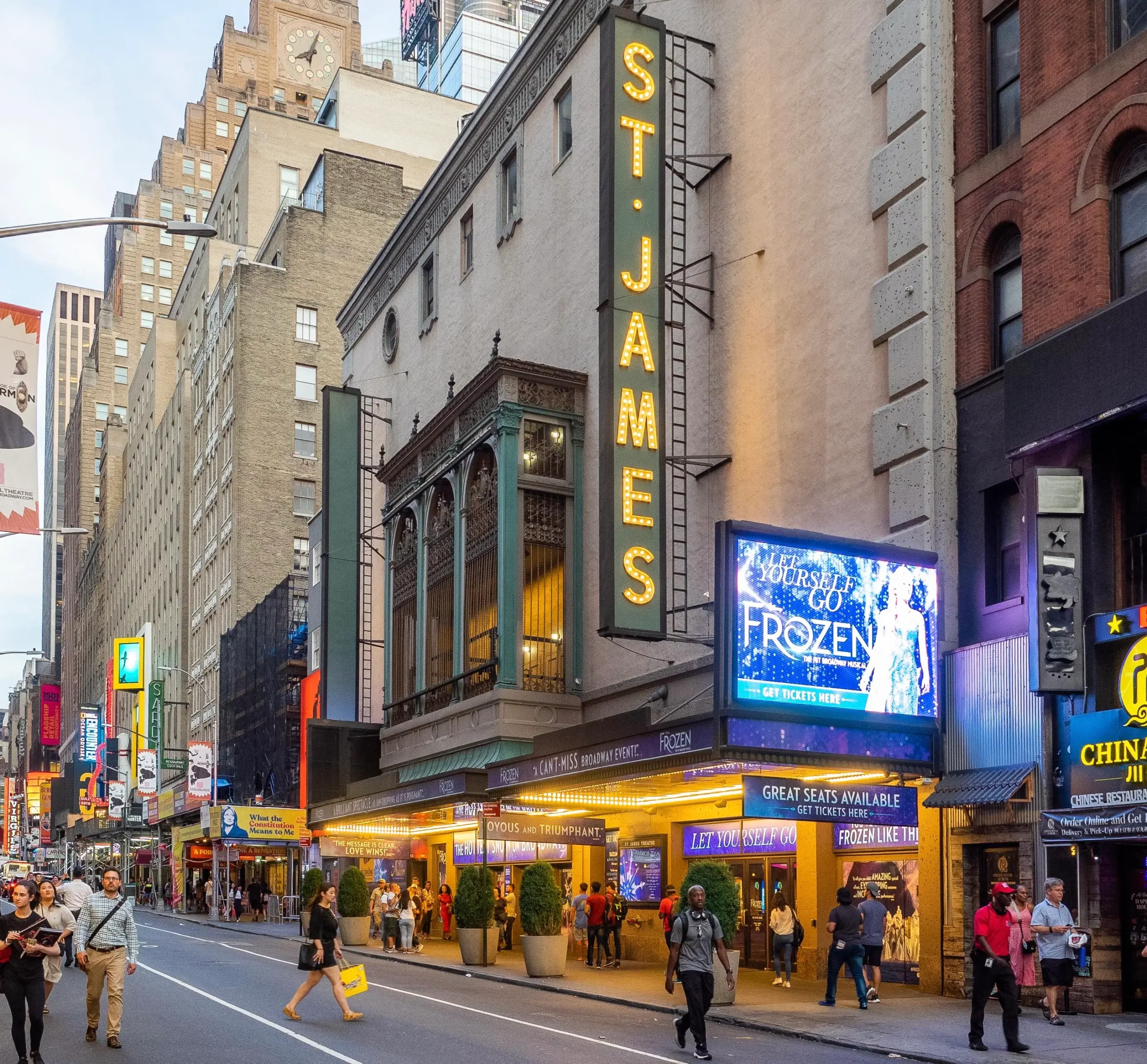 Street-level view of Broadway's historic St. James Theatre in New York City, featuring its illuminated vertical marquee, ornate façade, glowing entrance canopy, and digital Frozen billboard as pedestrians walk along West 44th Street in the Theater District.