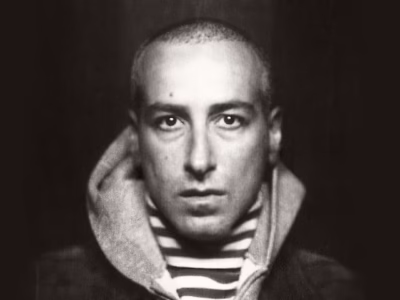 Black-and-white portrait of Francesco Risso looking directly into the camera with a closely shaved head and a serious expression. He wears a striped crewneck shirt layered beneath a gray hooded sweatshirt and a dark textured jacket. Dramatic lighting highlights his face against a nearly black background, creating a stark, minimalist editorial portrait