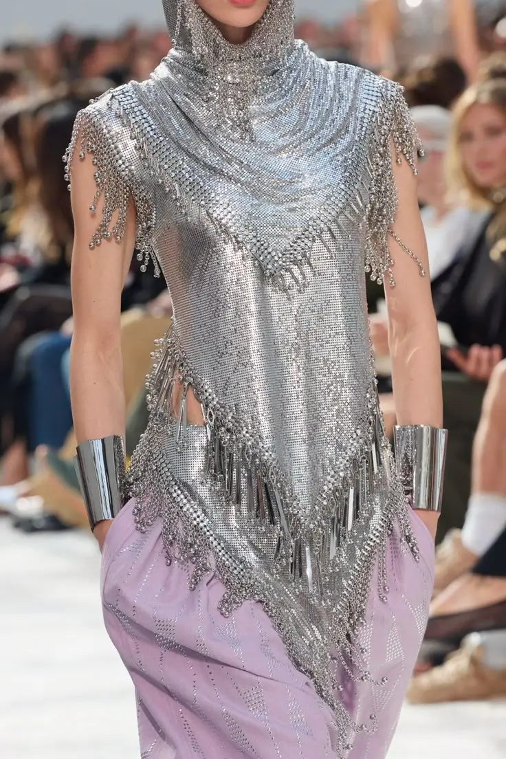 Close-up runway look featuring a shimmering silver chainmail hooded top with layered draping, crystal fringe, and metallic tassel detailing. The sleeveless garment is paired with pale lavender trousers embellished with subtle crystal patterns and finished with wide polished silver cuff bracelets. The blurred audience in the background emphasizes the intricate craftsmanship and reflective textures of the ensemble.