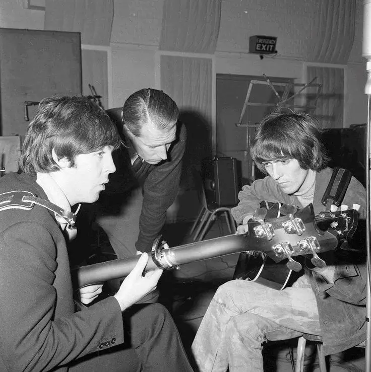 Black-and-white studio photograph of two Beatles examining a Gibson guitar with producer George Martin leaning in during the 1965 'Rubber Soul' recording sessions at EMI Studios in London.
