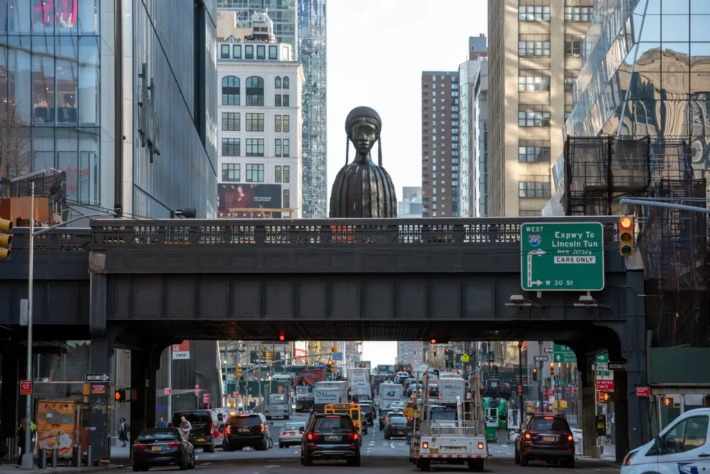 A wide street-level view beneath New York City's High Line shows steady traffic moving along a busy avenue lined with glass office towers and older brick buildings. Rising above the elevated park at the center is Simone Leigh's monumental bronze sculpture Brick House, depicting a woman with braided hair and a stylized cylindrical torso. An H&M storefront is view on the left, while a green highway sign directs drivers toward the Lincoln Tunnel via I-495 West. Late afternoon light reflects off surrounding skyscrapers, emphasizing the contrast between contemporary architecture, public art, and the city's constant flow of vehicle