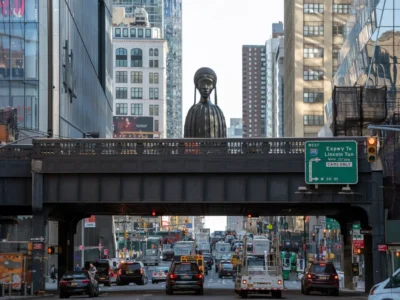 A wide street-level view beneath New York City's High Line shows steady traffic moving along a busy avenue lined with glass office towers and older brick buildings. Rising above the elevated park at the center is Simone Leigh's monumental bronze sculpture Brick House, depicting a woman with braided hair and a stylized cylindrical torso. An H&M storefront is view on the left, while a green highway sign directs drivers toward the Lincoln Tunnel via I-495 West. Late afternoon light reflects off surrounding skyscrapers, emphasizing the contrast between contemporary architecture, public art, and the city's constant flow of vehicle