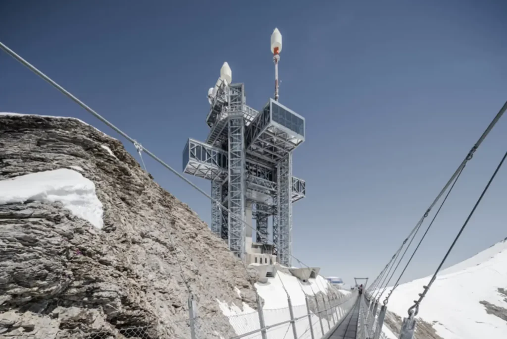 Modern steel observation tower with suspended glass lounges connected by a suspension bridge above a snowy alpine ridge