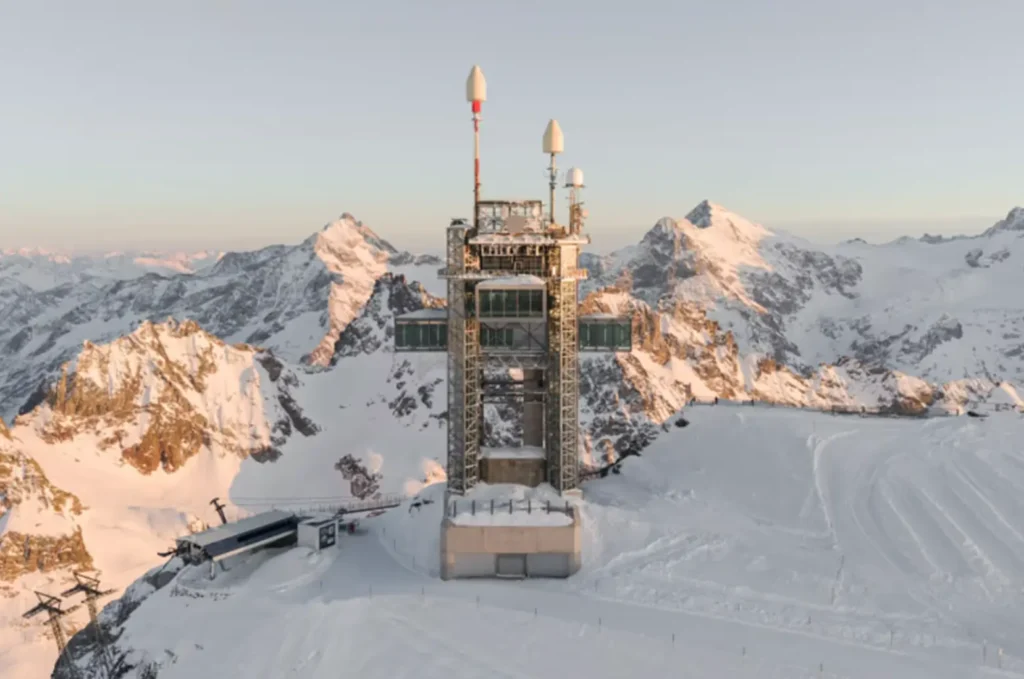 Modern steel observation tower with suspended glass lounges connected by a suspension bridge above a snowy alpine ridge