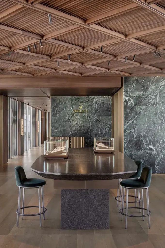 Elegant Rolex showroom featuring dark marble walls, walnut ceiling panels, glass display cases, and refined seating in a contemporary luxury interior