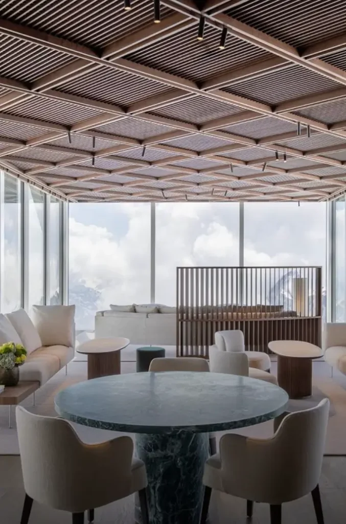Contemporary luxury lounge with marble table, plush seating, wood lattice ceiling, and panoramic alpine views through expansive glass wall