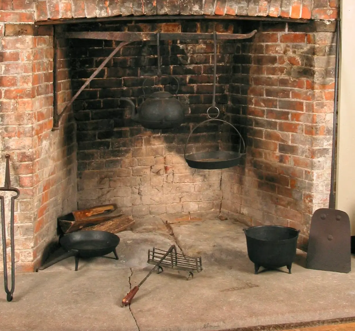 Colonial-era brick hearth fireplace with an iron crane holding a cast-iron kettle and hanging pot over the fire area. Traditional cooking tools, including a skillet, cauldron, fire grate, wood logs, and hearth accessories, rest on the stone floor.