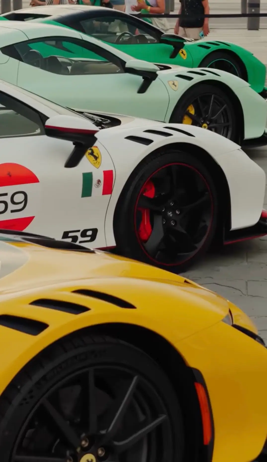 Lineup of Ferrari high-performance sports cars in yellow, white, mint green, and bright green finishes, showcasing sculpted front fenders, aerodynamic vents, black alloy wheels, and iconic Ferrari shield badges at an outdoor automotive event.