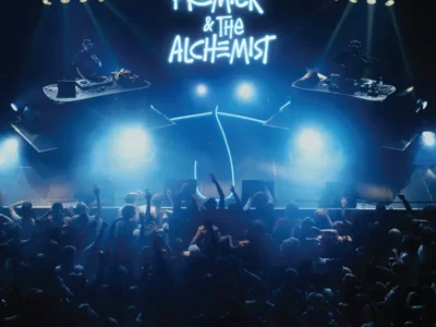 Concert stage featuring DJ Premier and The Alchemist performing from elevated DJ booths beneath a glowing neon backdrop, with bright blue stage lights illuminating a packed crowd below