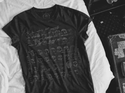 Black graphic Tee laid flat across a white bed, featuring a distressed monochrome print of repeated skull illustrations, handwritten script, and vertical text elements. A wooden nightstand with a glass, handwritten note, and ashtray sits beside the bed, while a worn suitcase and slippers complete the moody, vintage-inspired bedroom scene