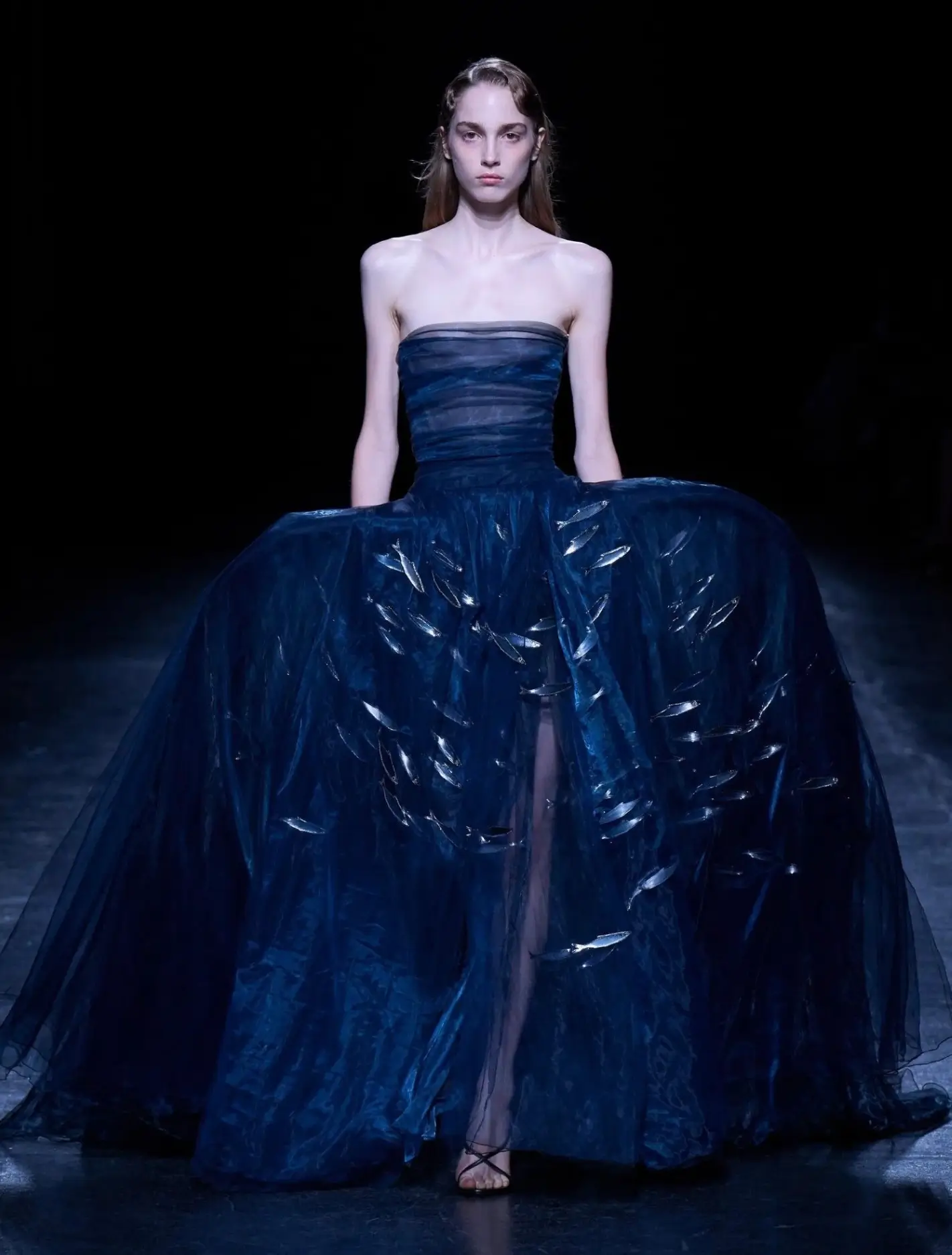 Model wearing a strapless deep navy evening gown with a sheer layered bodice, expansive sculptural skirt, and shimmering silver feather-like embellishments cascading across the front, showcased on a dark runway.