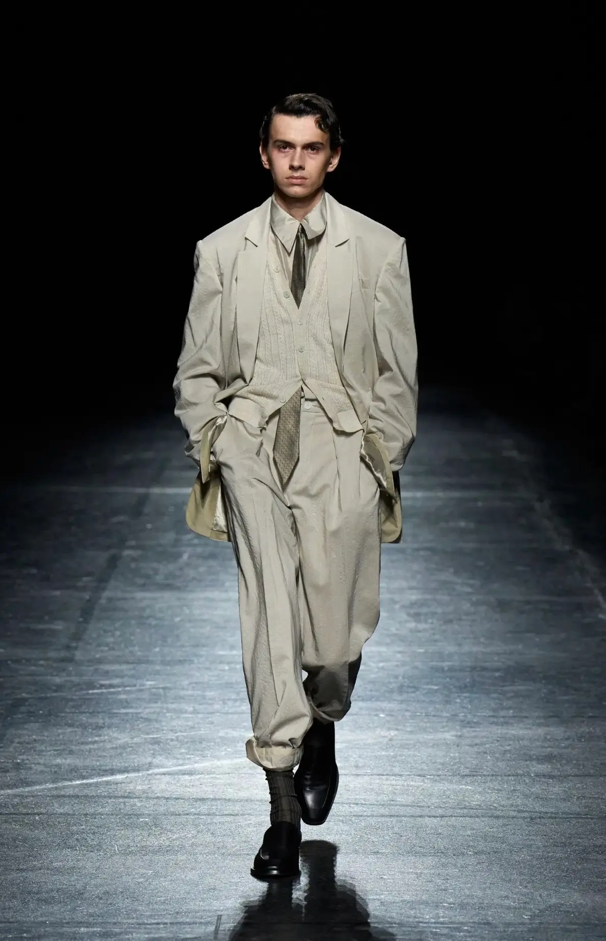 Model walking the runway in a relaxed ivory tailored suit featuring a softly structured blazer, textured knit vest, matching shirt and tie, pleated trousers, and black leather shoes with slouched socks.