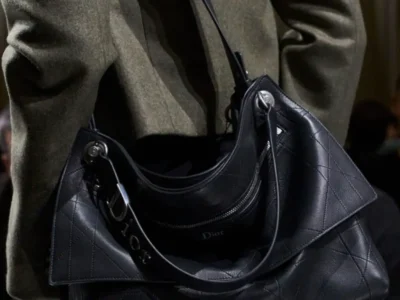 Black Dior leather shoulder bag showcasing an oversized quilted construction with a soft, slouching body, front zip pocket, detachable leather pouch, and polished silver hardware. Carried beneath an olive wool coat, the bag balances functional detailing with a clean, understated haute
