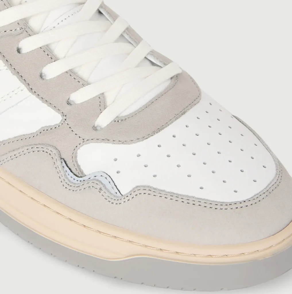 Close up of the sneaker's perforated white leather toe box, light gray suede mudguard, white flat laces, stitched overlays, and cream midsole