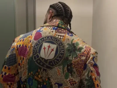 Review Scope Drake with braided hair wearing a richly patterned multicolor jacket featuring an oversized heraldic crest and embroidered Latin-style lettering across the back. The garment combines patchwork motifs, floral details, and geometric prints in an ornate, fashion-forward design