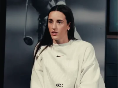 Caitlin Clark wearing an oversized white Nike shirt with a small black Swoosh and Giannis logo sits during an interview, with a black-and-white golf-themed backdrop behind them