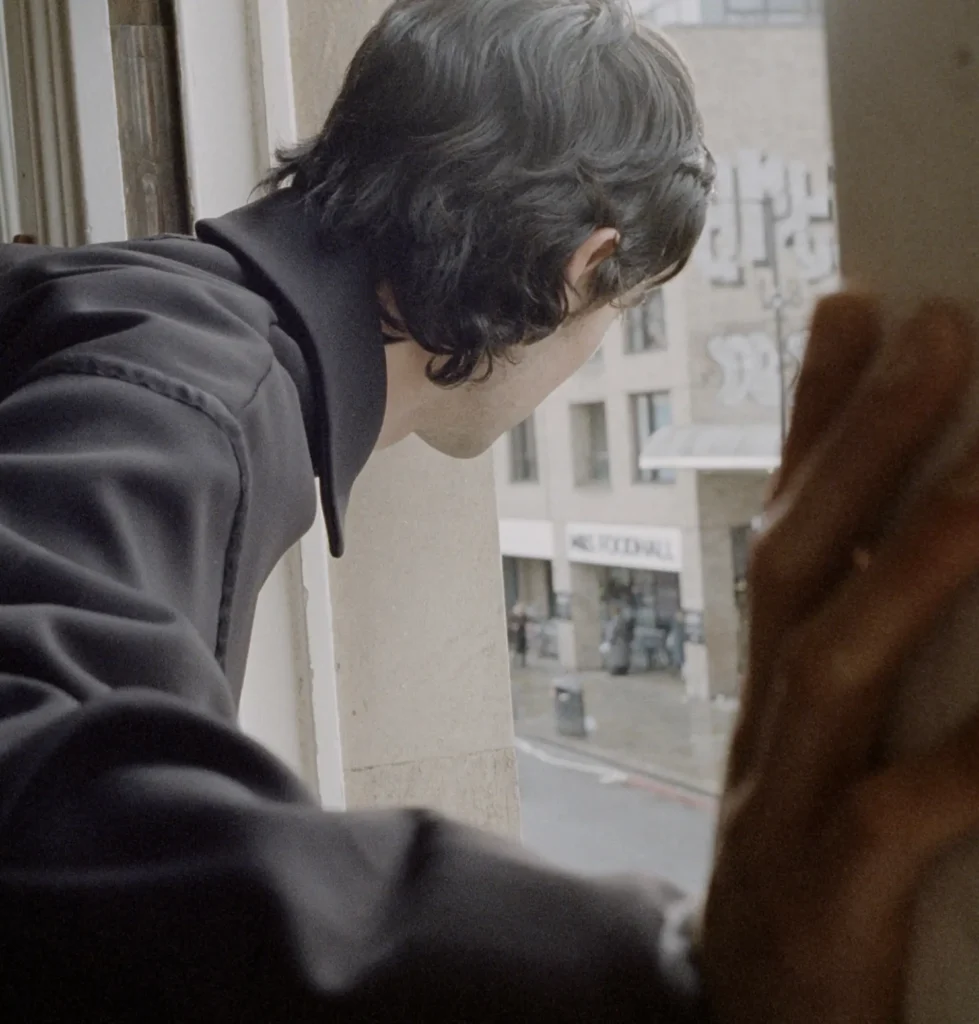 Male model in a structured black jacket leaning through an open upper-floor window and looking toward the street below, with another figure softly blurred in the foreground