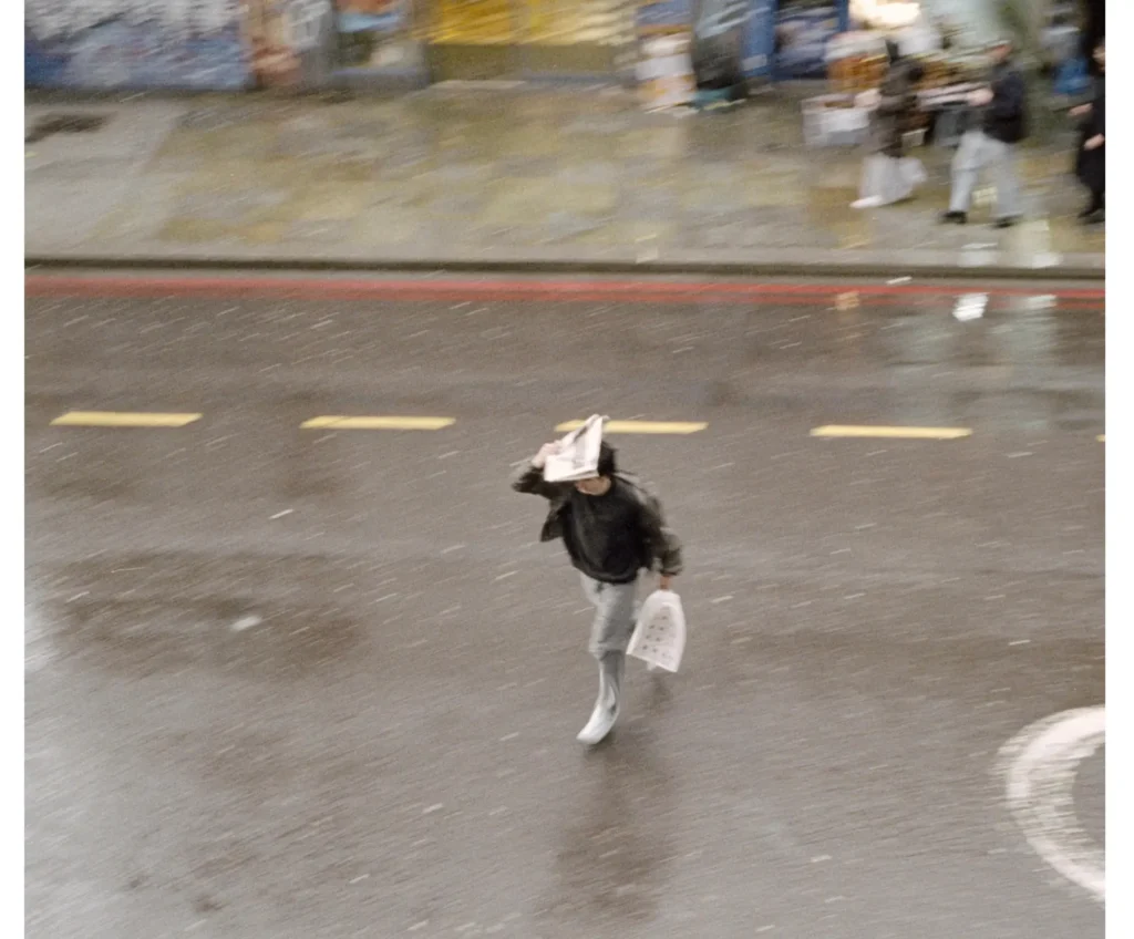 Blurred overhead street photograph of a pedestrian crossing wet traffic lanes while carrying shopping bags and shielding their head from the rain
