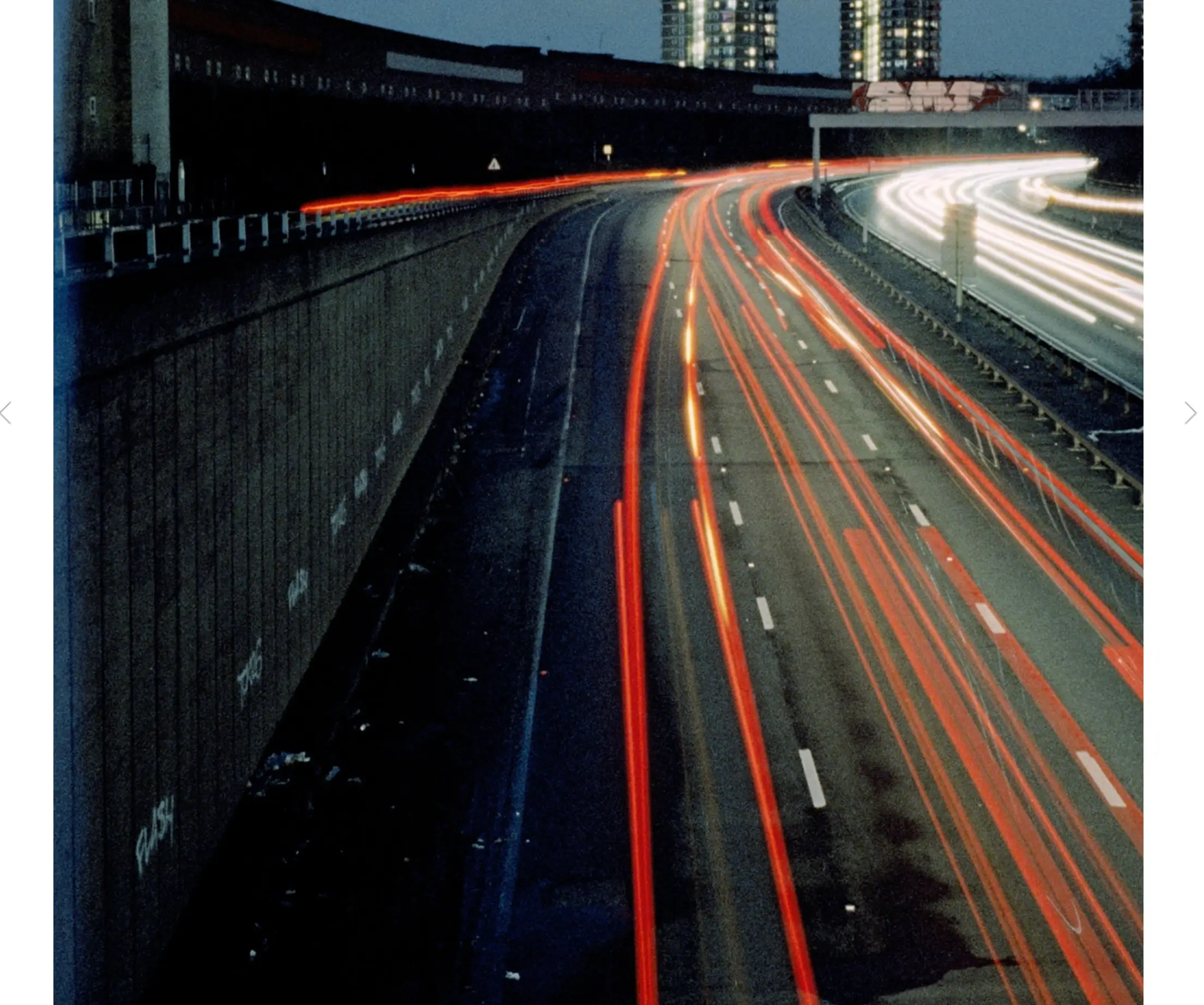 Long-exposure nighttime photograph of a multi-lane urban motorway, with red and white vehicle light trails curving through the roadway beneath illuminated residential towers.