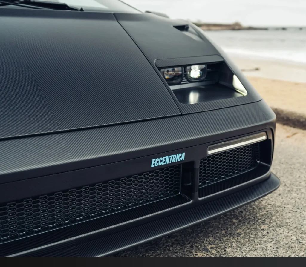 Front close-up highlighting the exposed projector headlight housing, carbon fiber hood texture, honeycomb grille, and show Eccentrica badge