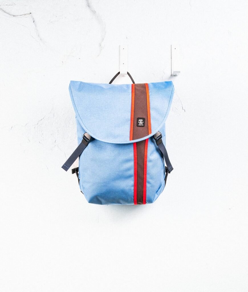 Light blue Crumpler backpack hanging against a white textured wall, featuring a flap closure with contrasting vertical stripe detailing and adjustable side strap
