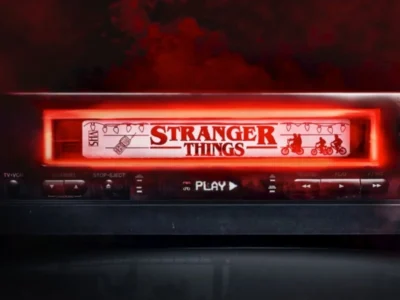 Close-up of a retro VHS player with a glowing red cassette labeled "Stranger Things" inserted, illuminated by a dramatic crimson light against a dark, smoky background. The VCR's front panel displays playback controls and a lit "PLAY" indicator, evoking the nostalgic horror aesthetic of the series