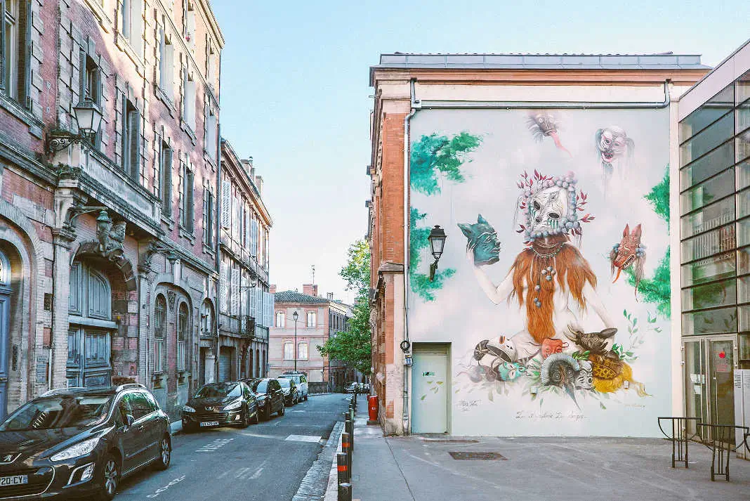 Street art mural by Miss Van in Toulouse, France, painted on the side of a building, depicting a masked figure adorned with beaded headpieces and animal masks, surrounded by illustrated hybrid creatures, shown above a narrow cobblestone street lined with parked cars and traditional brick buildings.