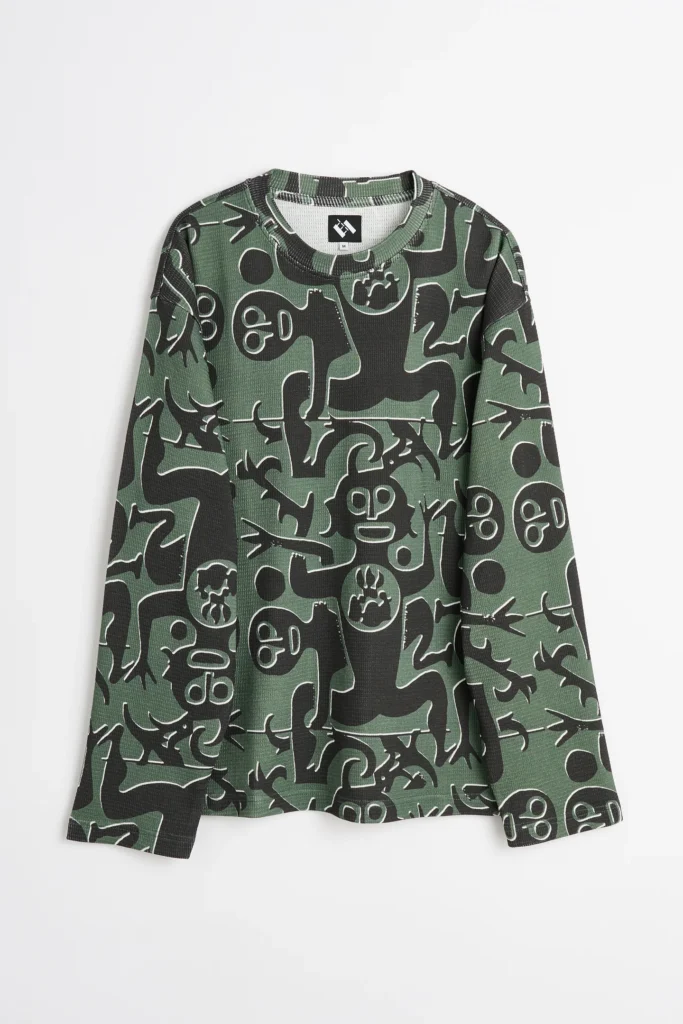 A green waffle knit long sleeve shirt covered in an all over abstract print of black cartoon inspired figures, faces, arrows, and organic shapes. The playful pattern spans the entire garment against a neutral white backdrop