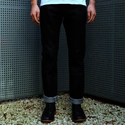 Front view of dark indigo selvedge denim jeans with cuffed hems, worn with black leather boots and photographed against a perforated metal wall over white gravel.