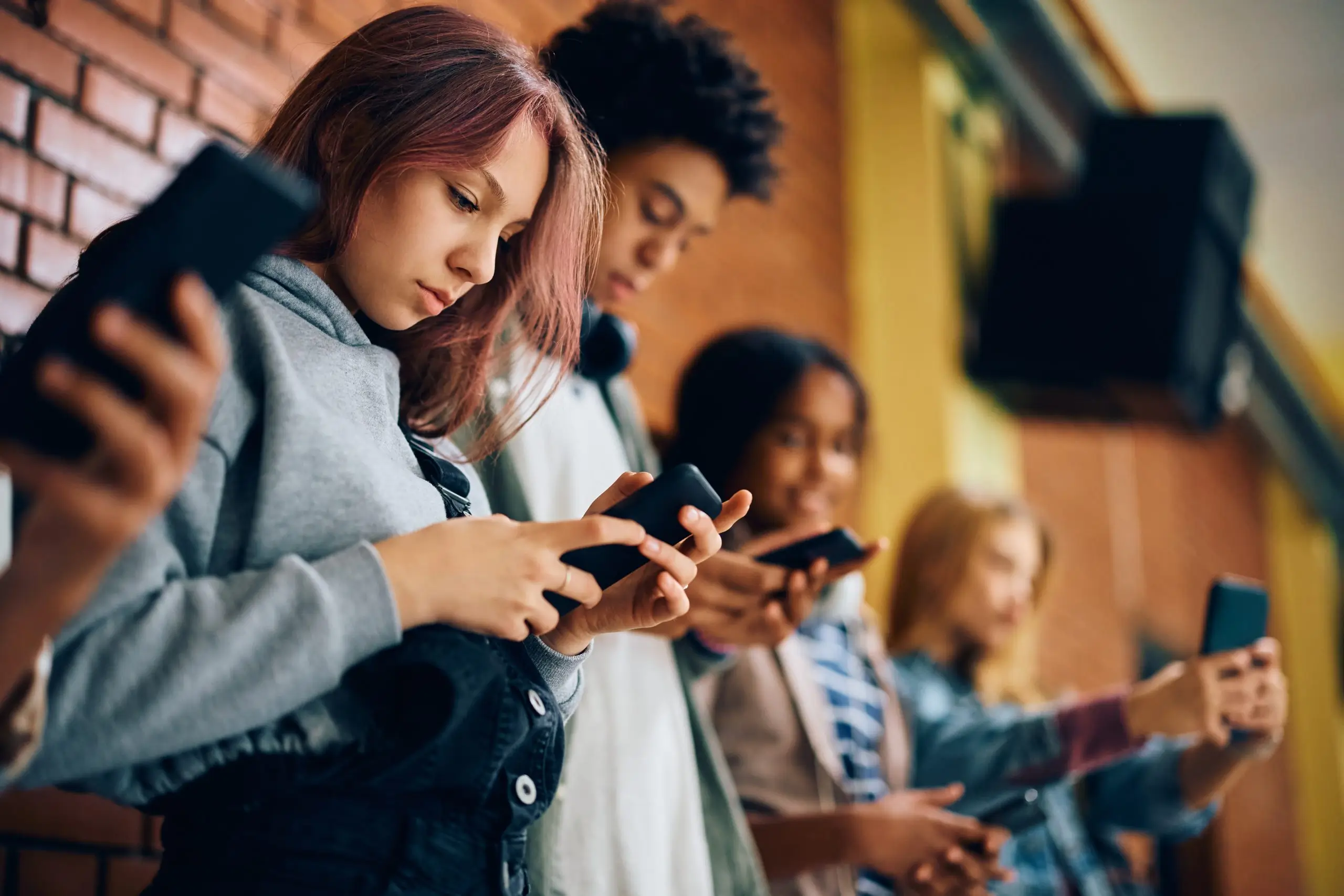 Teenagers using smartphones together outside a school building.