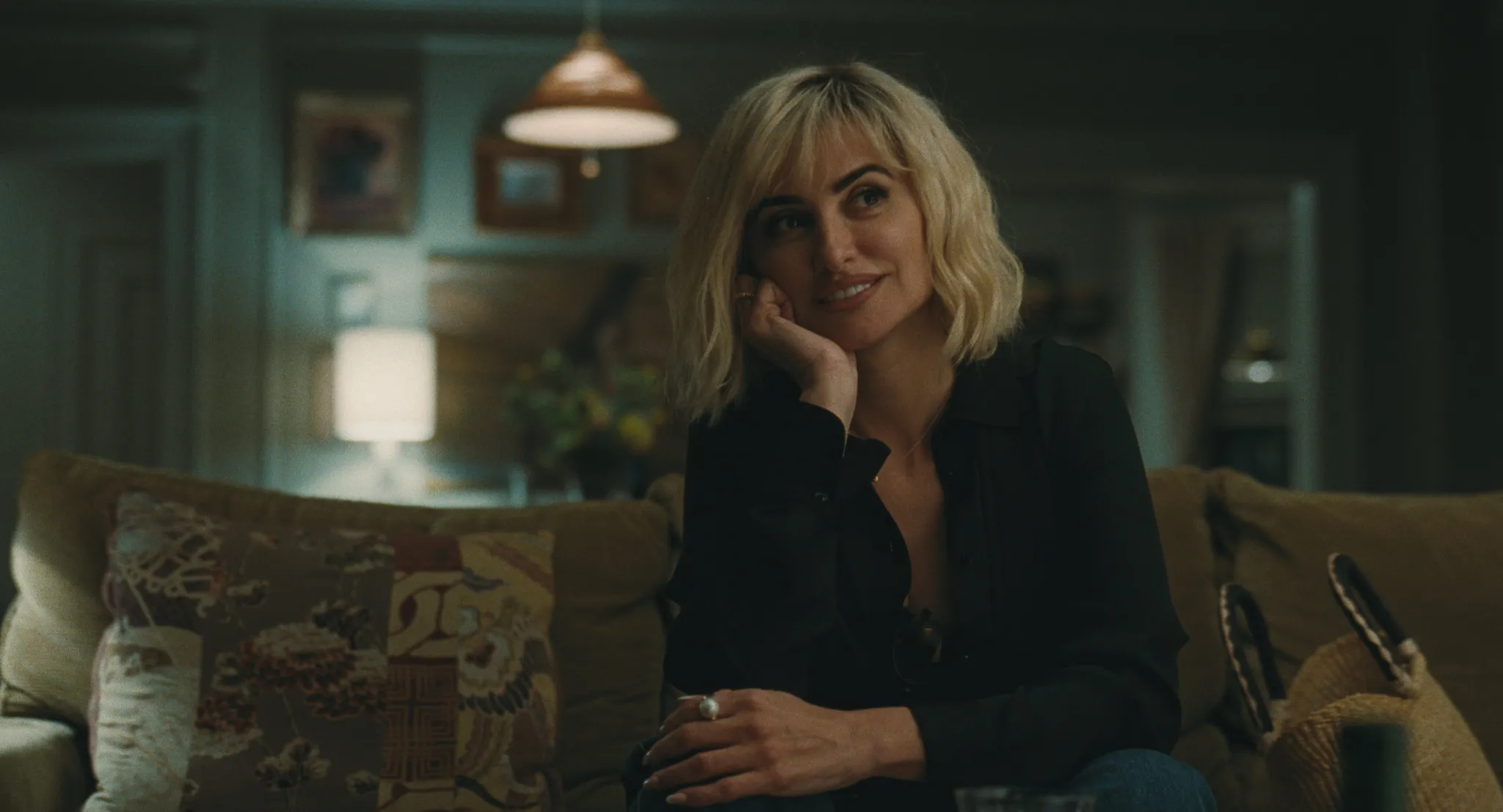 Penélope Cruz sits on a living room sofa with a warm smile, wearing a black blouse and blonde bob hairstyle in a softly lit scene from The Invite.