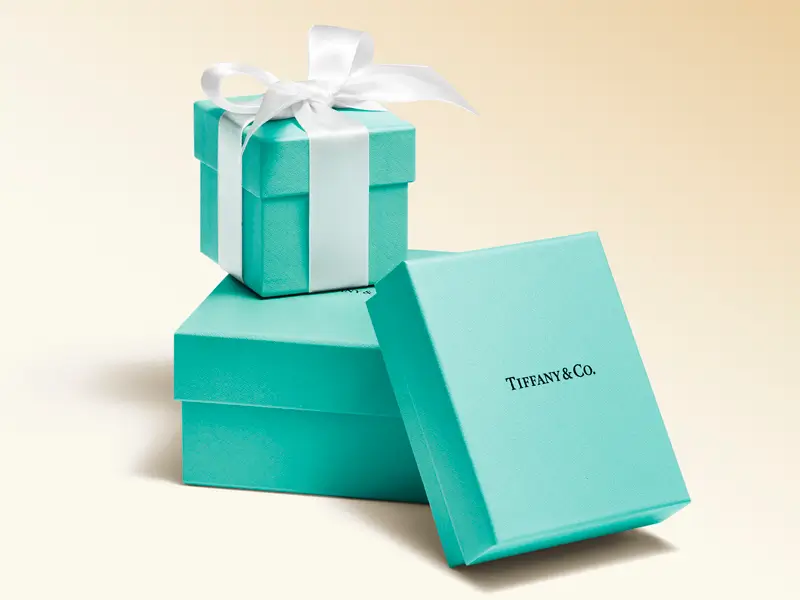 Stack of Tiffany & Co.'s signature robin's egg blue gift boxes, including a small box wrapped with a white satin ribbon and a larger presentation box with its lid leaning against the side, photographed on a soft cream background.