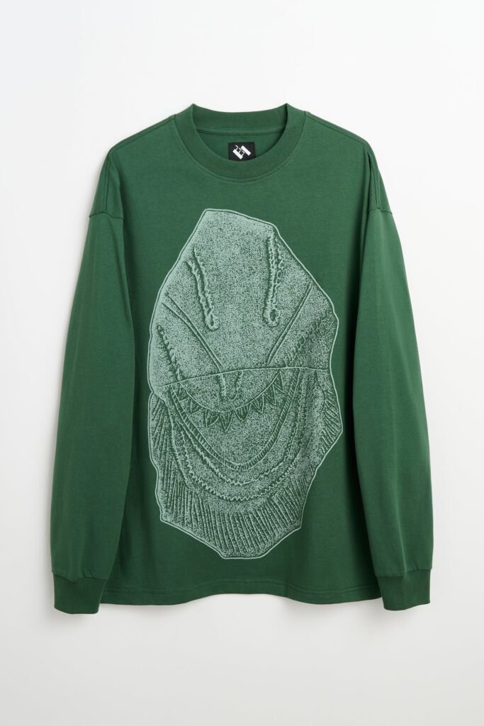 A forest green long sleeve T shirt featuring a large white line illustration of a hooded, toothy creature centered on the front. Displayed flat on a white background, the graphic combines horror inspired imagery with hand drawn texture