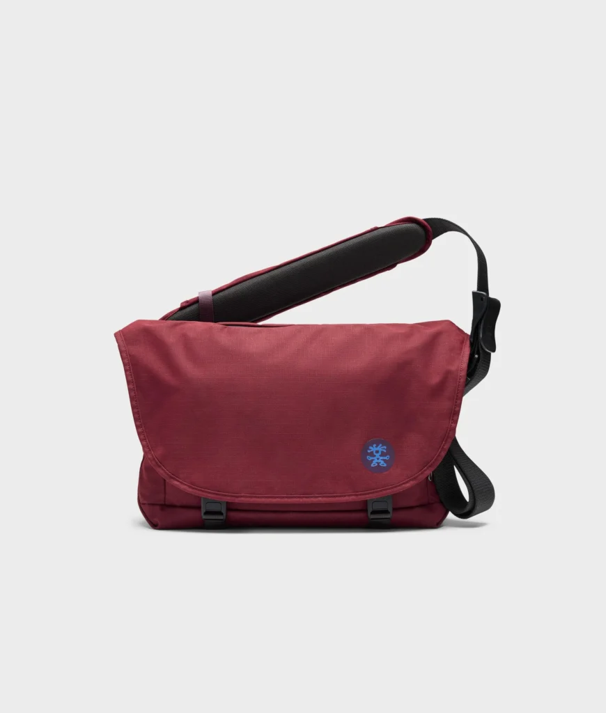 Burgundy Crumpler messenger bag displayed against a clean white background, featuring a wide flap closure, padded shoulder strap, black hardware, and a contrasting circular logo badge