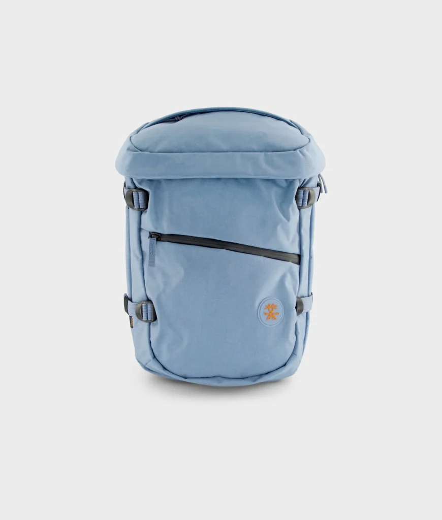 Light blue Crumpler backpack displayed against a clean white background with a curved top profile, angled front zip pocket, side compression straps, and a circular logo patch
