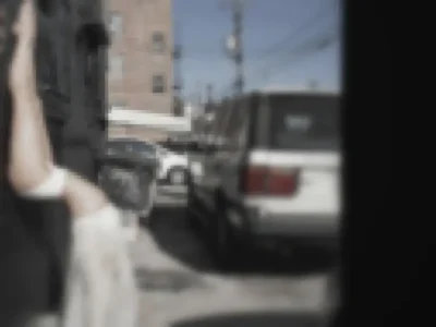 Blurry street photo shot from a doorway: Babe America raised arm and light garment in the foreground, a parked white SUV, brick apartment building, utility poles, and clear sky behind
