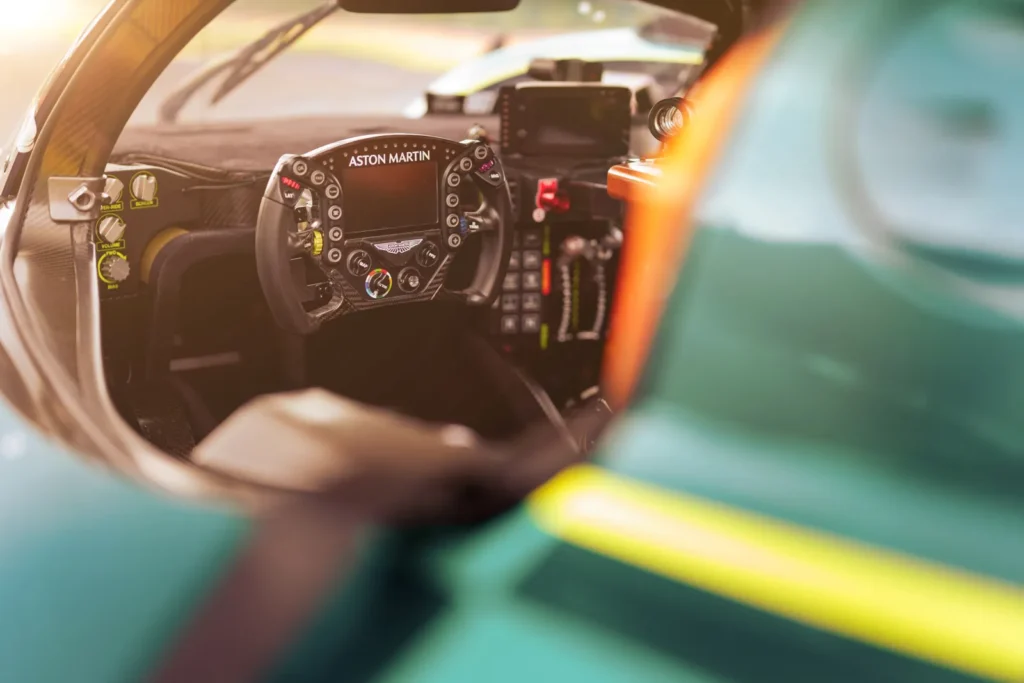 Interior cockpit view of an Aston Martin race car showcasing the Formula-style steering wheel, digital instrument display, carbon-fiber dashboard, racing controls, and minimalist driver-focused cabin illuminated by warm natural light