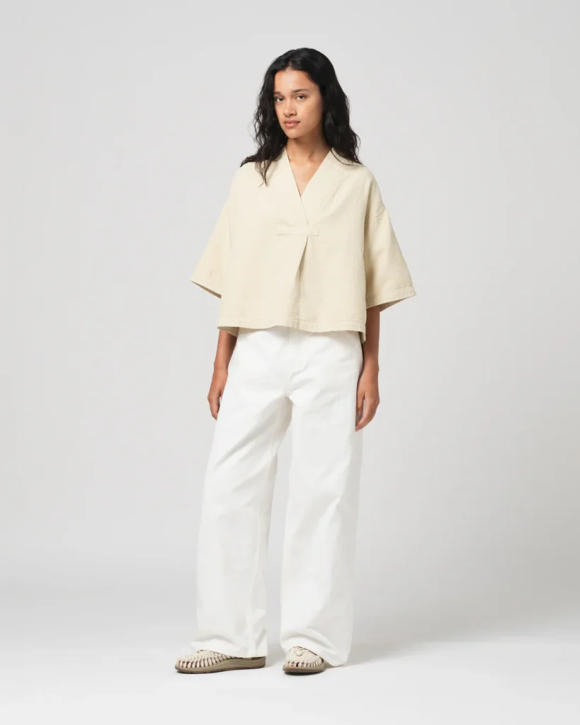 Model wearing a cropped cream wrap-style top with wide sleeves, paired with loose white trousers and woven slip-on sandal