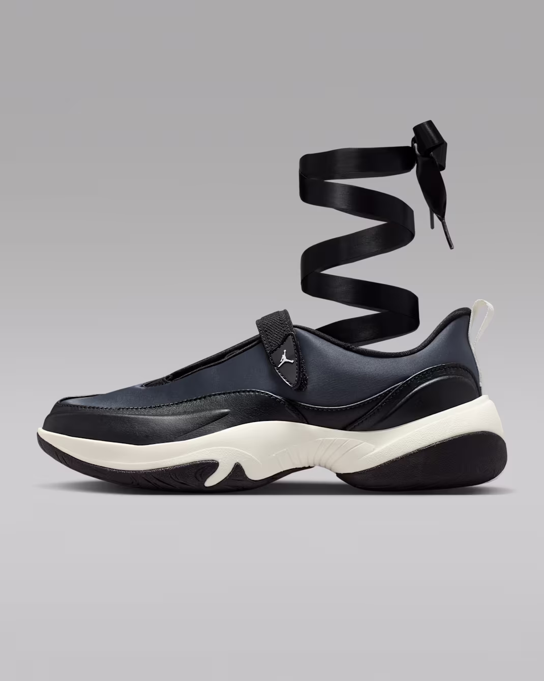 Black and white Jordan Pointe slip-on sneaker with a sculpted chunky sole, minimalist leather upper, adjustable midfoot strap with Jumpman logo, elongated wraparound ribbon lace, and contrasting white heel pull tab.