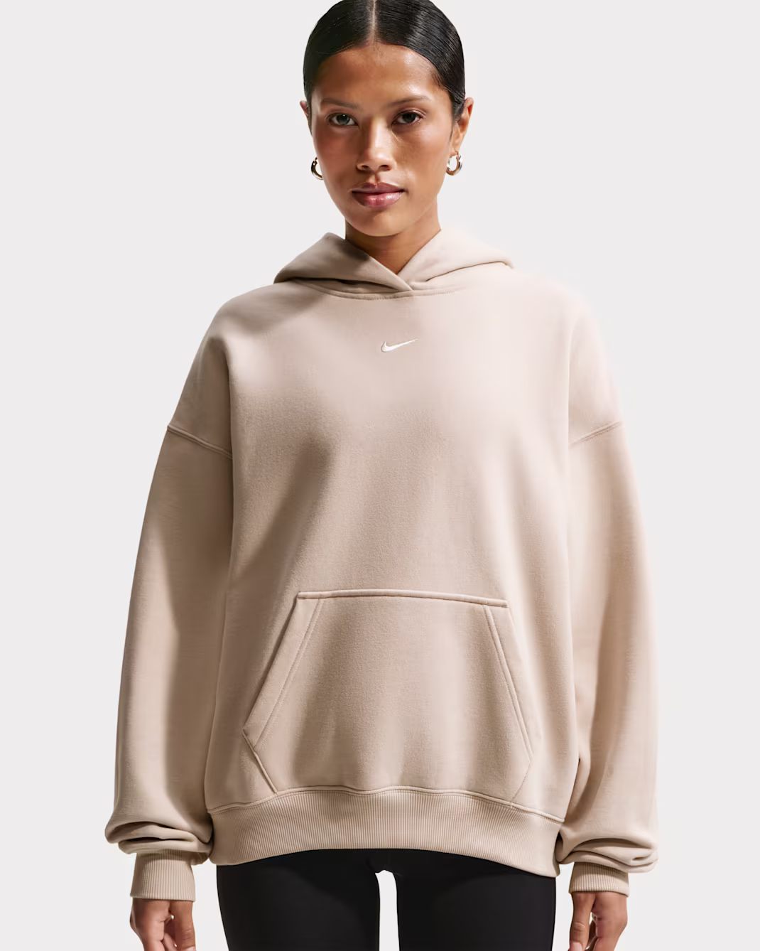 Front view of the oversized Nike Sportswear fleece hoodie in a soft beige color, featuring a relaxed fit, adjustable hood, kangaroo pocket, ribbed cuffs and hem, and an embroidered white Swoosh logo at the chest.