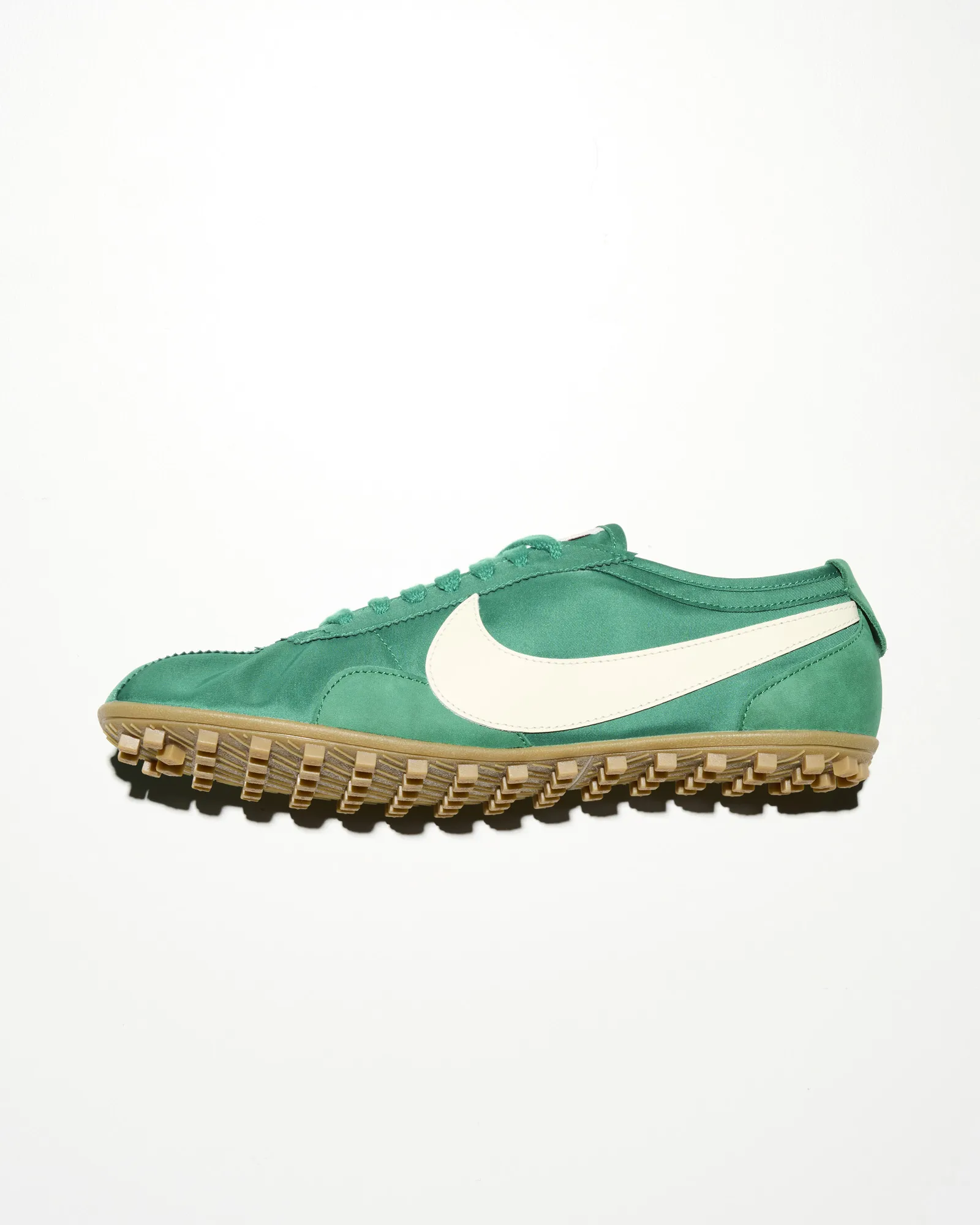 Side profile of a green vintage-inspired Nike training shoe with a white leather Swoosh, textured gum rubber lug outsole, and low-cut silhouette, photographed against a clean white studio background.
