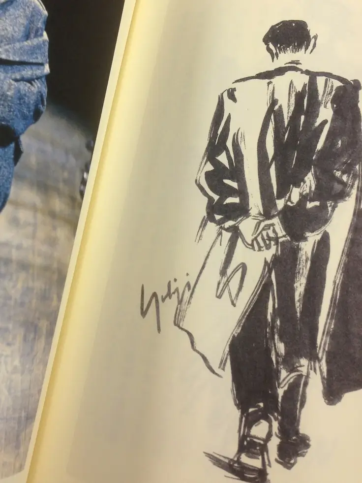Expressive black ink fashion sketch of a figure viewed from behind, wearing a long flowing coat with hands clasped behind the back, drawn on a cream book page.