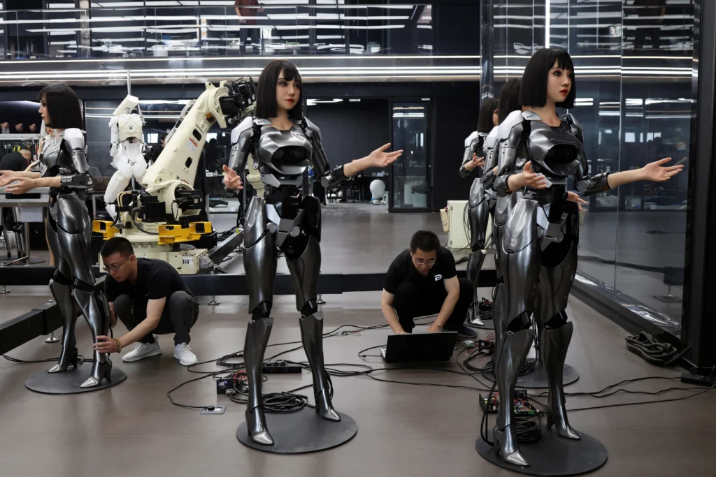 White service robot holding a small flower in a demonstration area, equipped with a large camera-like eye and articulated robotic arms. Additional robots stand behind it, highlighting hospitality and customer service applications in a clean exhibition set