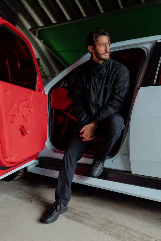 Alpine Formula 1 driver Pierre Gasly sitting on the open door sill of the Alpine A290 Rallye, wearing a black Alpine x Lacoste tracksuit beside the red-trimmed interior