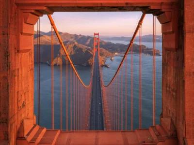 Golden Gate Bridge suspension tower framing the bridge roadway and Marin Headlands at sunrise, with warm orange light illuminating the steel structure, suspension cables, and calm waters of San Francisco Bay
