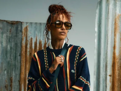 Portrait of a Alewya wearing oversized square sunglasses and a navy striped knit sweater with colorful vertical bands, standing before weathered corrugated metal panels in an industrial fashion editorial