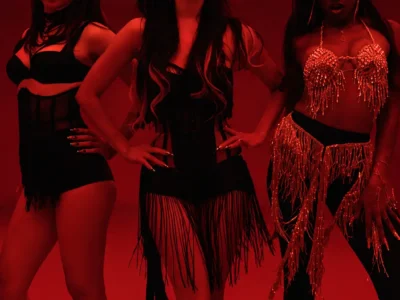 Katseye group pose mid-dance beneath deep red lighting, wearing black fringed stage outfits with their hands positioned at their hip