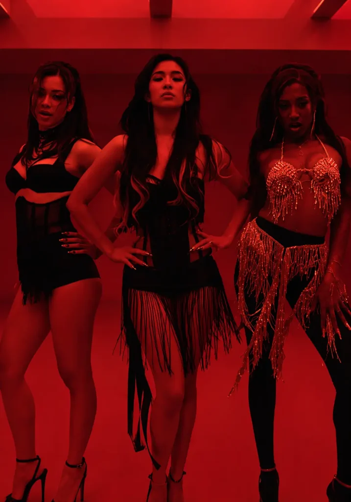 Katseye group pose mid-dance beneath deep red lighting, wearing black fringed stage outfits with their hands positioned at their hip
